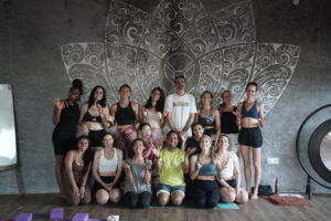 Diverse group of people smiling and waving in a yoga studio with a mandala wall.